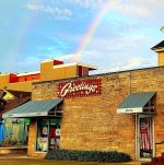 Storefront of Greetings Gift Shop in Bloomington, Indiana, with a rainbow arching over the building against a partly cloudy sky