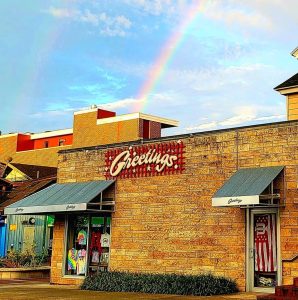 Storefront of Greetings Gift Shop in Bloomington, Indiana, with a rainbow arching over the building against a partly cloudy sky