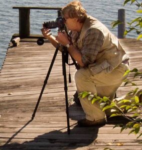 Photographer looking through a camera mounted on a tripod while standing on a wooden dock beside the water