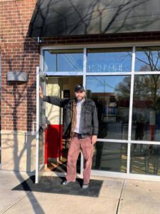 Travers Marks, owner of Max's Place in Bloomington, Indiana, standing in the doorway of the cafe on a sunny day. The brick exterior and glass door reflect a welcoming atmosphere.
