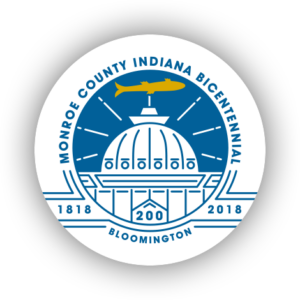 A circular logo for the Monroe County Indiana Bicentennial (1818-2018), featuring a blue and white illustration of the Monroe County Courthouse dome, surrounded by radiating lines. A golden fish is positioned above the dome. The words "Monroe County Indiana Bicentennial" encircle the top, and "Bloomington" appears at the bottom.