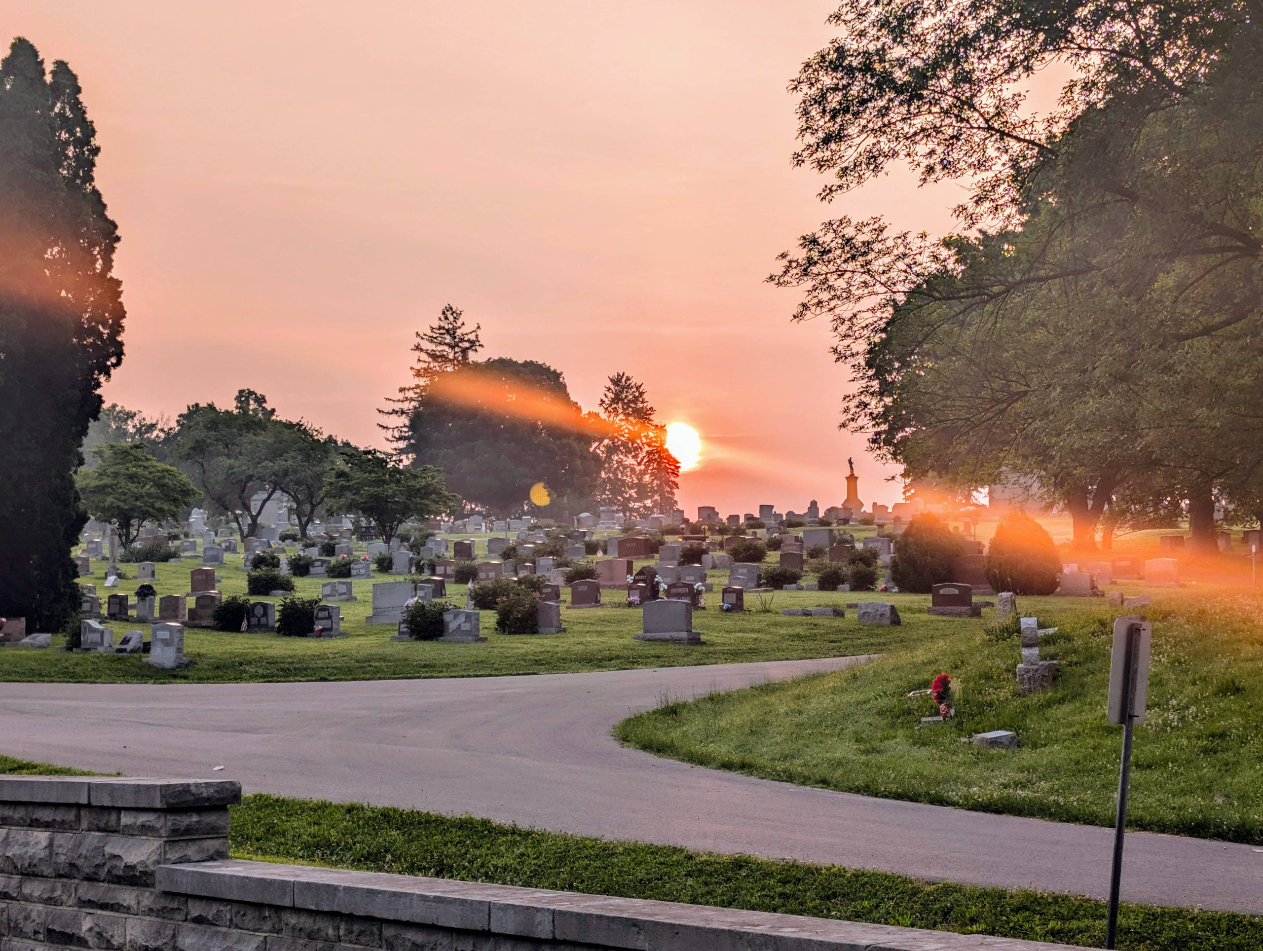 Sunrise over Rose Hill Cemetery with gravestones, large trees, and warm orange lens flare across the tranquil morning scene.