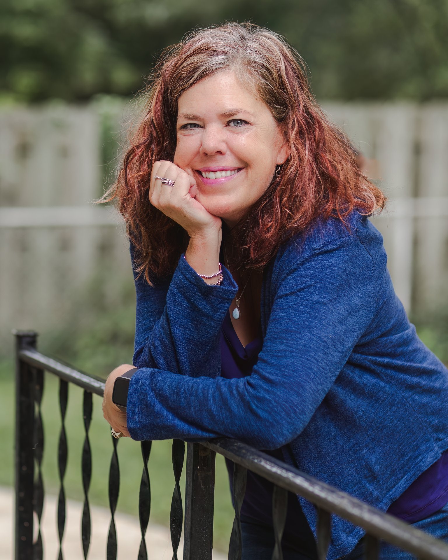 Colleen Wells, writer and creative writing facilitator, smiling warmly while leaning on a wrought iron railing, wearing a blue cardigan. Outdoor portrait with soft green background.