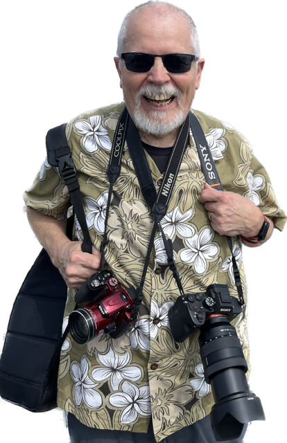 Doc Searls smiling while wearing a Hawaiian shirt and carrying multiple cameras, including Sony and Nikon models, at a local Bloomington event