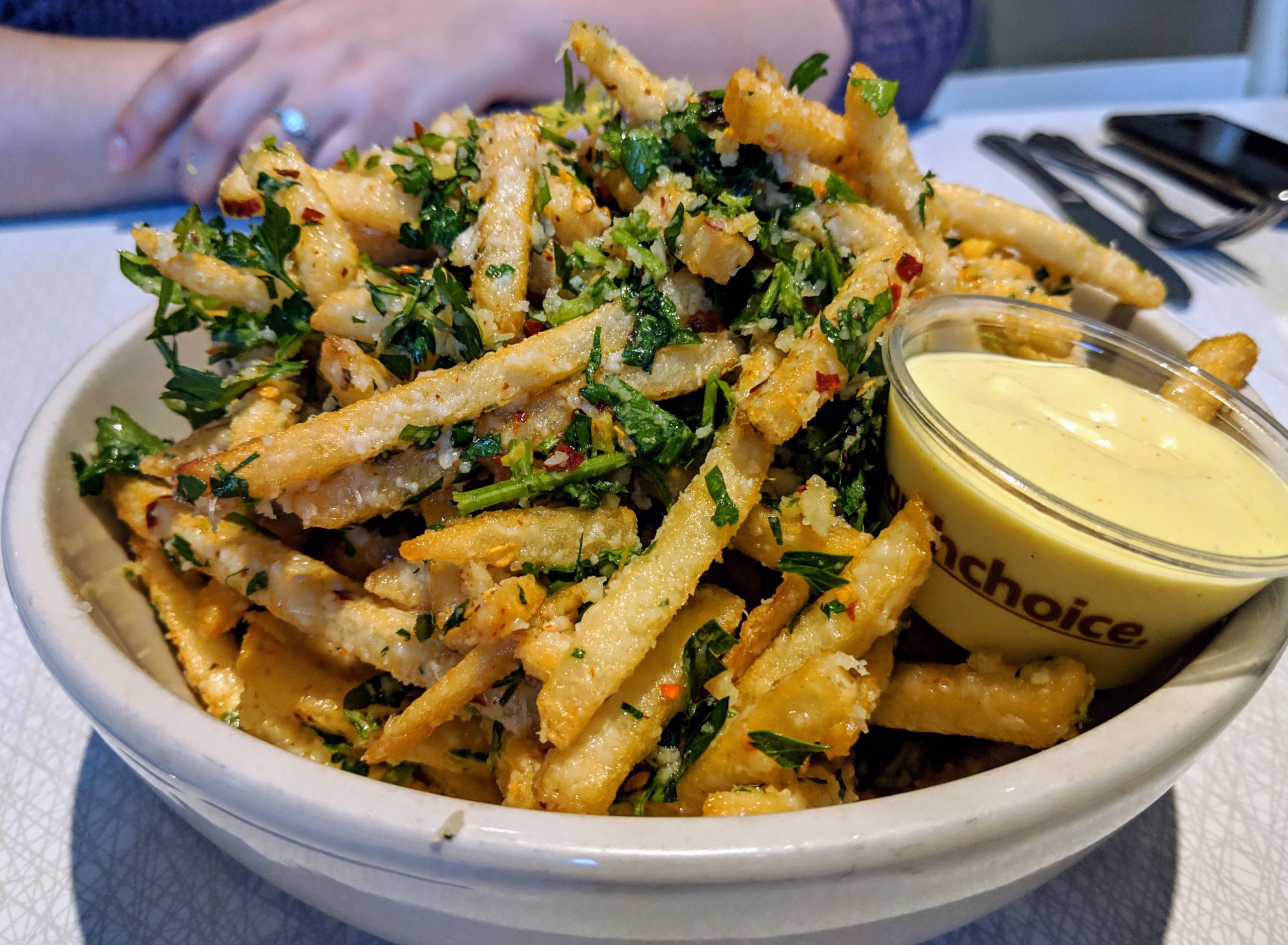 A bowl of crispy garlic herb Parmesan fries garnished with parsley and red pepper flakes, served with a side of creamy aioli dipping sauce