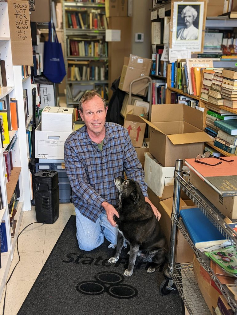 Joe Grant, owner of Academic Scholarly Books, kneeling beside a black dog in a used academic bookstore filled with shelves, boxes, and non-fiction titles