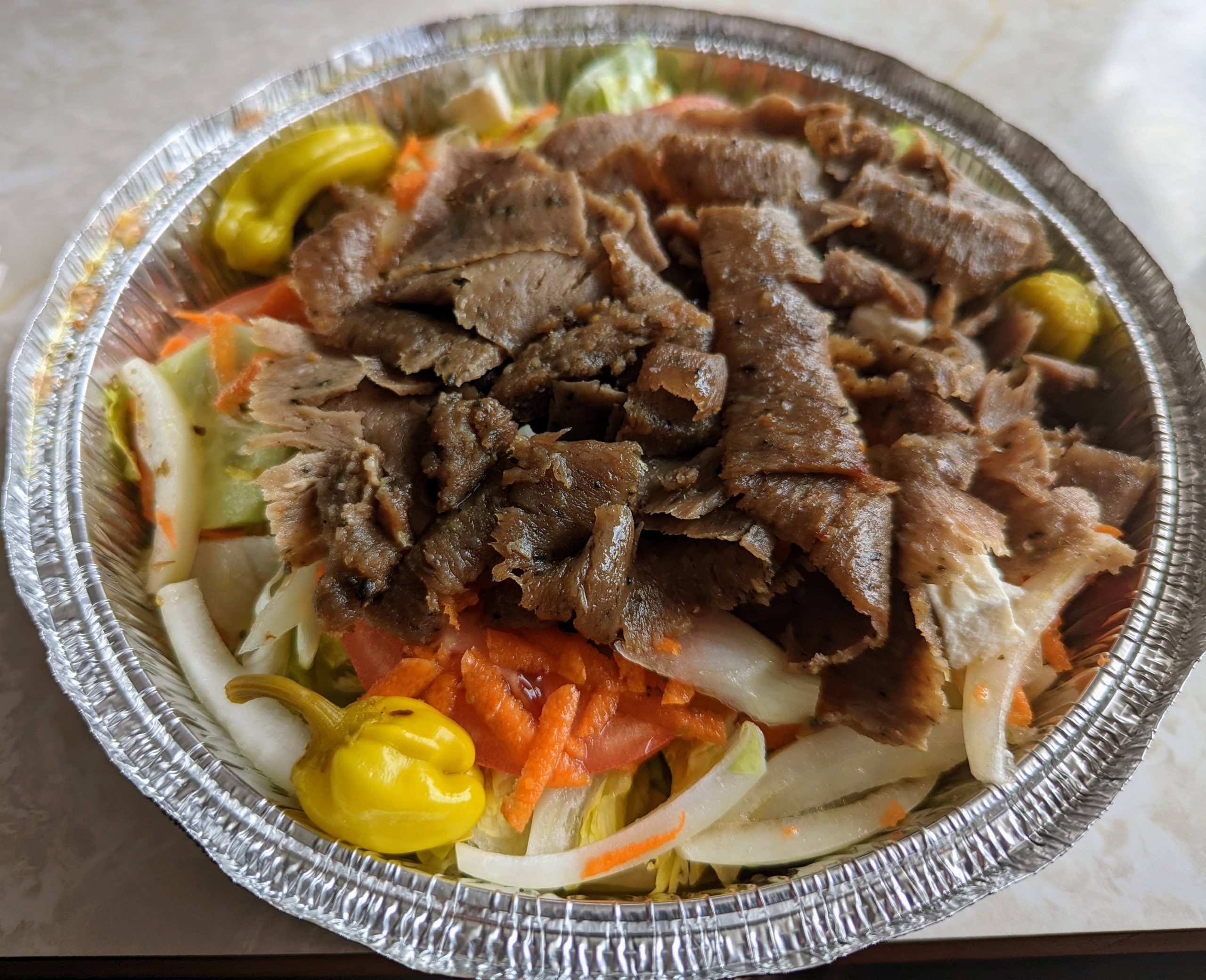 Gyro salad from King Gyros in Bloomington, featuring shaved lamb gyro meat over fresh lettuce, carrots, onions, tomato slices, and banana peppers in a foil container.
