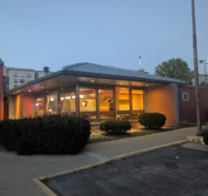 La Charreada at dusk: the glass-walled dining room under a slate-blue hip roof, lit orange and yellow, with orange diagonal siding at the ends