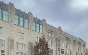 A long limestone campus building with tall windows beside a street, under a cloudy sky