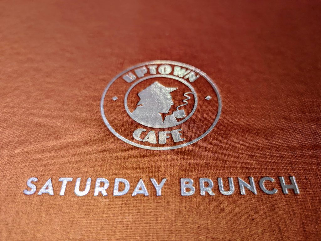 Close-up of Uptown Cafe's Saturday Brunch menu cover with silver embossed logo and text on a textured brown background